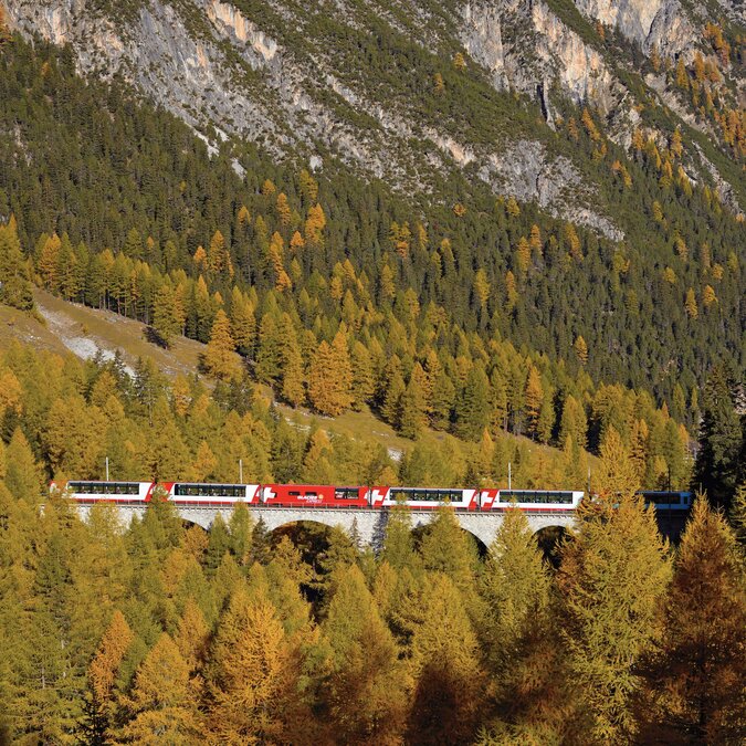 Der Glacier Express fährt über ein Viadukt durch herbstlich gefärbte Wälder im Albulatal, umgeben von hohen Bergflanken | © Glacier Express
