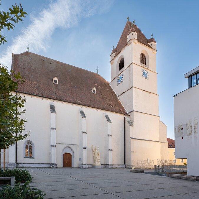 Der Dom St. Martin in Eisenstadt mit seinem markanten Kirchturm und der schlichten historischen Fassade. Der Vorplatz wird von Bäumen und moderner Architektur umgeben, während der blaue Himmel die Szene prägt. | © Eisenstadt(c)Burgenland Tourismus_Andreas Hafenscher (45)
