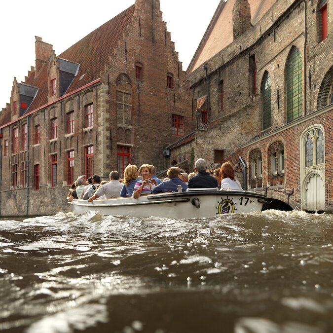 Boot mit Besuchern auf dem Kanal in Brügge, umgeben von mittelalterlichen Backsteinhäusern. | ©  Visit Brugge – Bootsfahrt