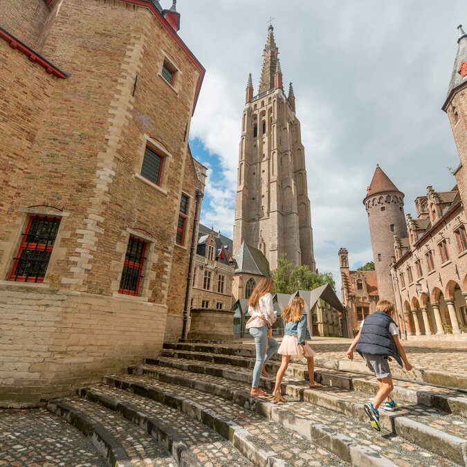Mehrere Personen gehen eine breite Steintreppe in der Altstadt von Brügge hinauf. Umgeben sind sie von historischen Backsteinbauten, darunter ein hoher Kirchturm und weitere mittelalterliche Gebäudeteile. Der Platz ist von Kopfsteinpflaster, Bögen und charakteristischer Architektur geprägt. | © Jan Darthet 