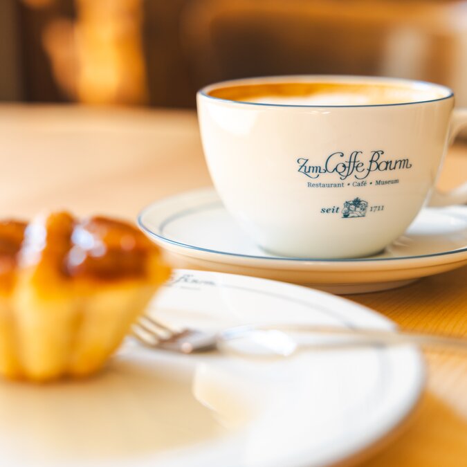 Kaffeetasse mit Logo des Coffe Baum Leipzig und Gebäck auf Holztisch im warmen Licht. | © Philipp Kirschner | Leipzig Travel