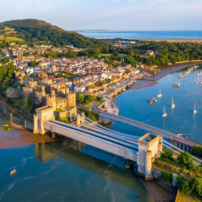 Luftaufnahme von Conwy in Wales mit mittelalterlicher Burg, Brücke, Hafen und Küstenstadt vor gruener Landschaft und ruhigem Meer. | © shutterstock 2203832967
