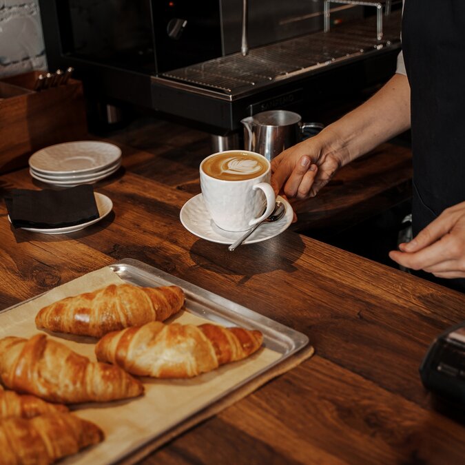 Ein Barista platziert einen Cappuccino mit Latte Art neben frisch gebackenen Croissants auf einer Holztheke in einem gemütlichen Café-Ambiente | © Shutterstock 2393949781