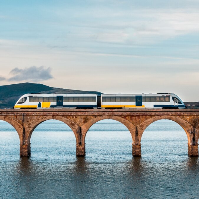 Zugreise durch Spanien mit modernem Zug auf historischer Steinbrücke über Wasser, weiter Landschaft und ruhiger Stimmung unter hellem Himmel. | © shutterstock 1378378817